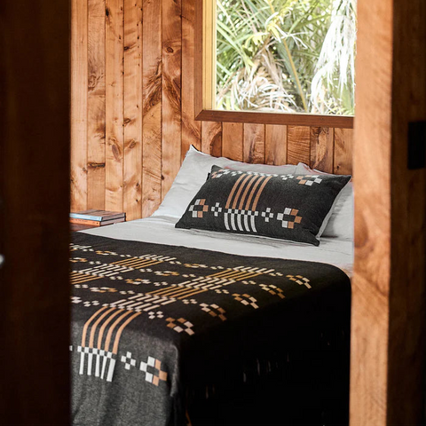 Bed with patterned blanket and pillow in a wooden room with a window showing greenery