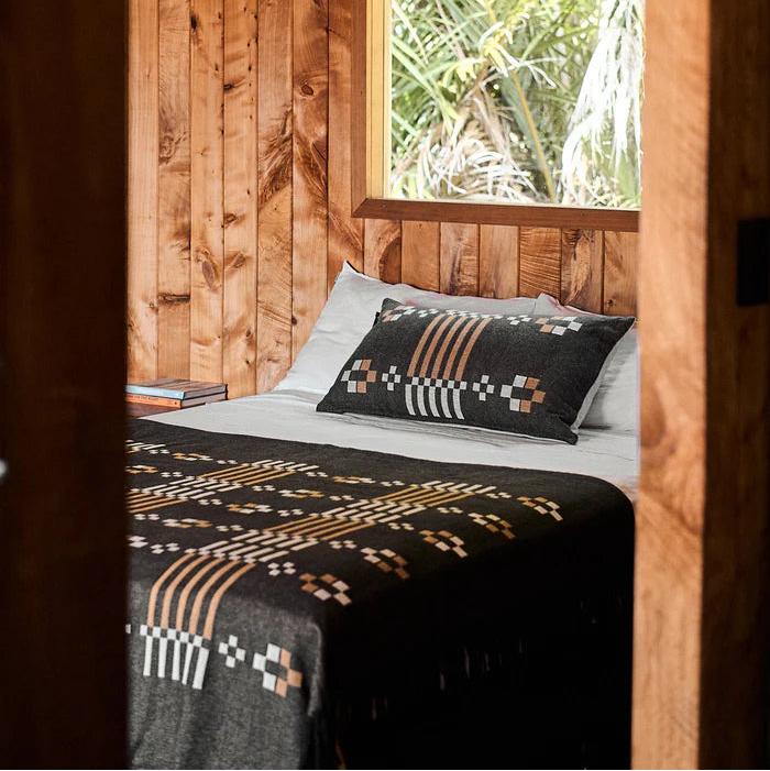 Bed with patterned blanket and pillow in a wooden room with a window showing greenery