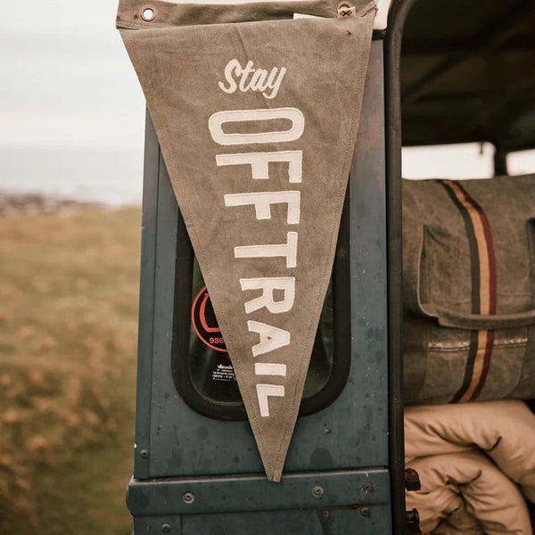 A triangular canvas Pony Rider camping pennant banner hanging from the side of a vintage green vehicle outdoors. The banner is a warm tan color with white text reading 'Stay OFF TRAIL' and is shown next to a sleeping bag inside the open vehicle door.