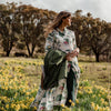 Woman standing in a field of yellow flowers wearing a Binny dress patterned with vintage vegetable images and holding a green jacket.