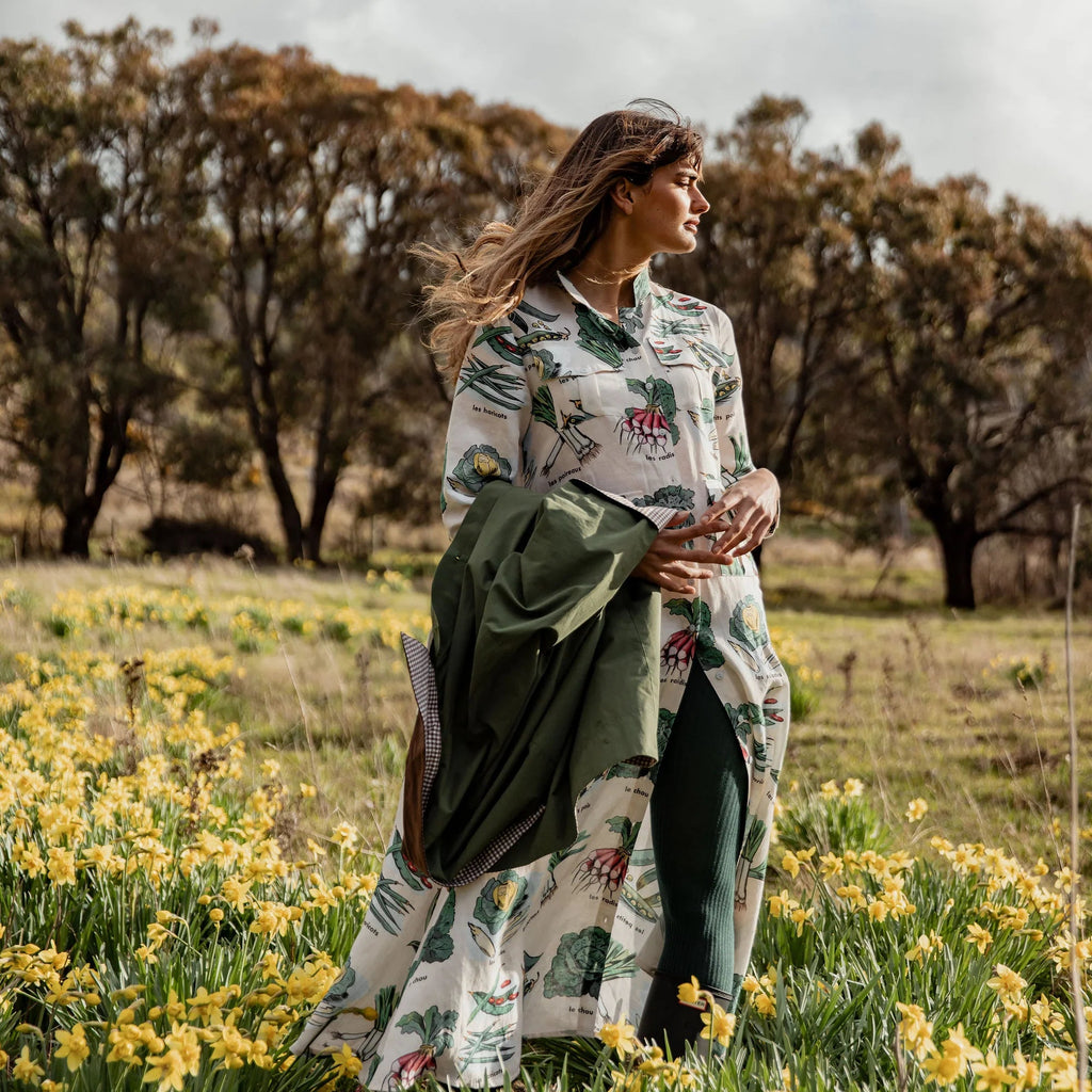 Woman standing in a field of yellow flowers wearing a Binny dress patterned with vintage vegetable images and holding a green jacket.