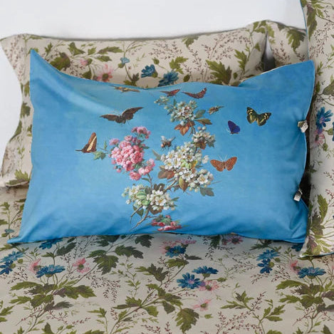 A rectangular blue decorative pillow with a colorful floral and butterfly print, styled on top of a matching floral-patterned bedspread and pillowcase.