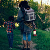 Two people walking on a path in a forest with one carrying a lantern and the other wearing a backpack.