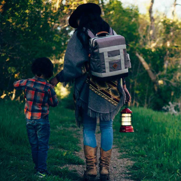 Two people walking on a path in a forest with one carrying a lantern and the other wearing a backpack.