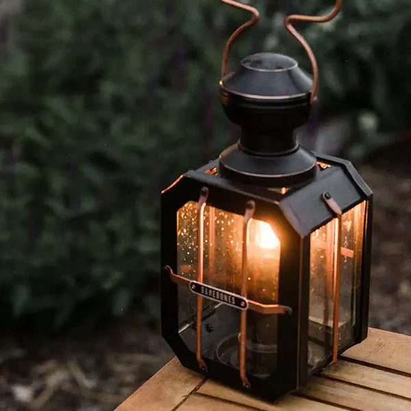 Barebones Decorative lantern with a warm glow on a wooden surface outdoors.