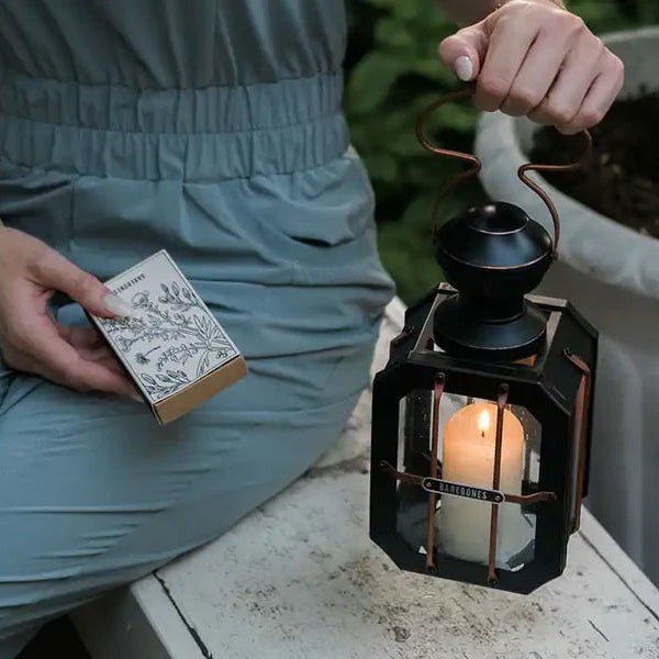 Person holding a Barebones lantern with a lit candle and a small book outdoors.