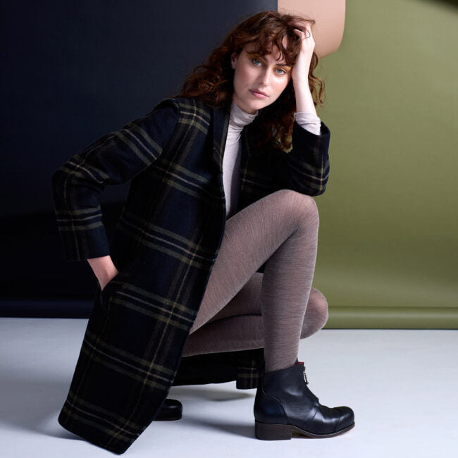 Woman in a plaid coat and black boots sitting on a bench against a green wall.