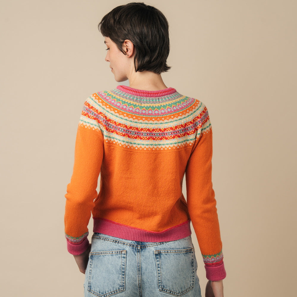 Person wearing an orange Eribe sweater with a colorful pattern and blue jeans on a beige background