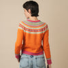Person wearing an orange Eribe sweater with a colorful pattern and blue jeans on a beige background