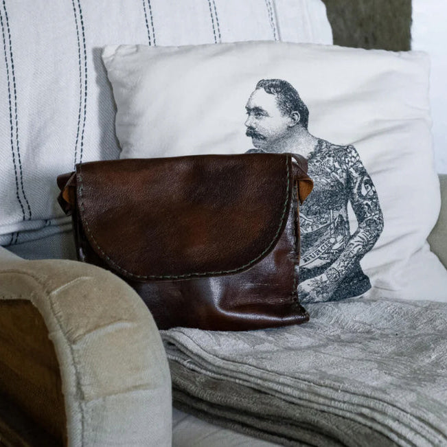 Brown leather bag on a couch next to a pillow with a tattooed man design.