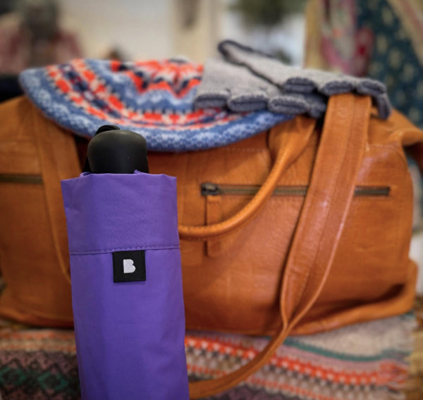 Purple umbrella with a Blunt logo in front of a brown leather bag on a colorful surface.