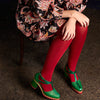 Person wearing a patterned dress, red tightology tights, and green shoes on a dark background