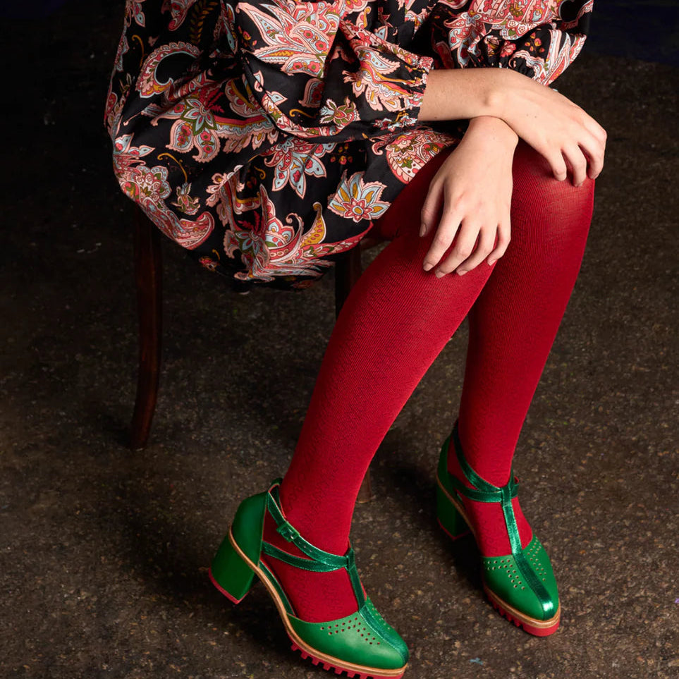 Person wearing a patterned dress, red tightology tights, and green shoes on a dark background
