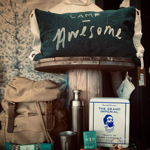 Camp-themed pillow with 'Camp Awesome' text, backpack, thermos, and other items on a wooden stand.