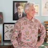 Man wearing a pink floral shirt sitting in a room with light blue walls and framed pictures.