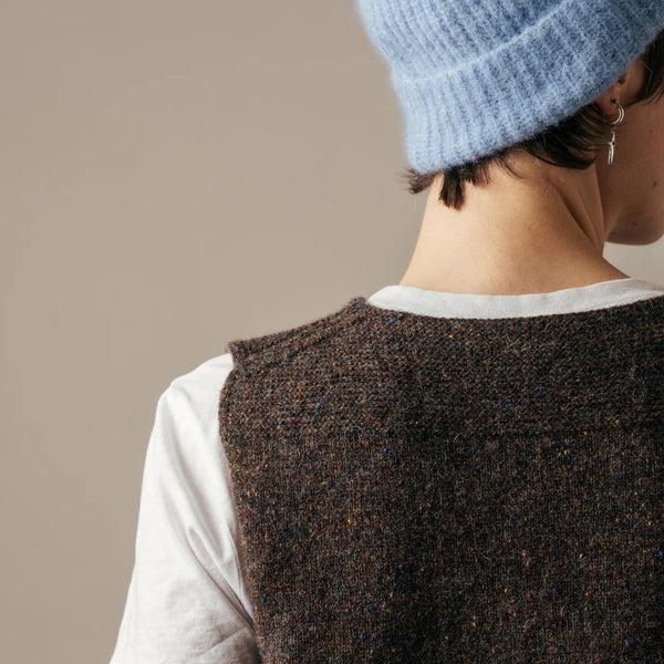 Person wearing a brown knitted vest over a white shirt with a blue knit hat on a neutral background