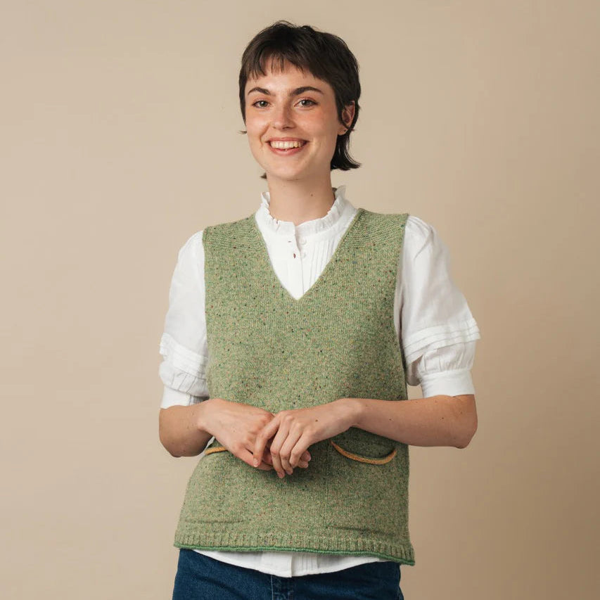 Person wearing a green knitted vest over a white shirt with blue pants on a beige background