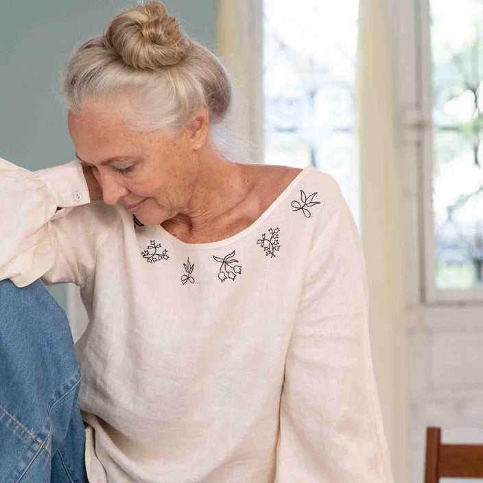 Woman wearing a white blouse with floral embroidery sitting on a wooden stool indoors.