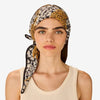 A woman wearing a patterned Inoui Editions headscarf with a black, white, and mustard yellow daisy-and-spotted leopard design, paired with a cream ribbed tank top, against a light gray background.