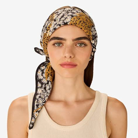 A woman wearing a patterned Inoui Editions headscarf with a black, white, and mustard yellow daisy-and-spotted leopard design, paired with a cream ribbed tank top, against a light gray background.