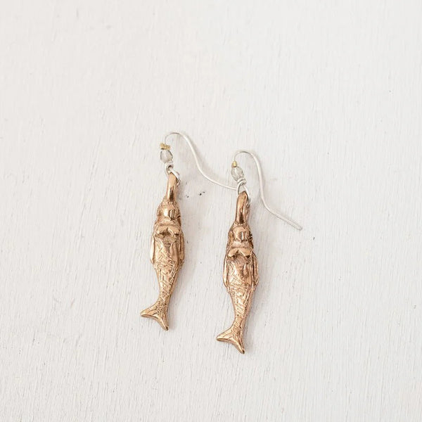 A pair of mermaid shaped dangle earrings in a textured bronze finish, suspended from simple silver-toned fishhook ear wires, displayed against a clean, off-white textured background.