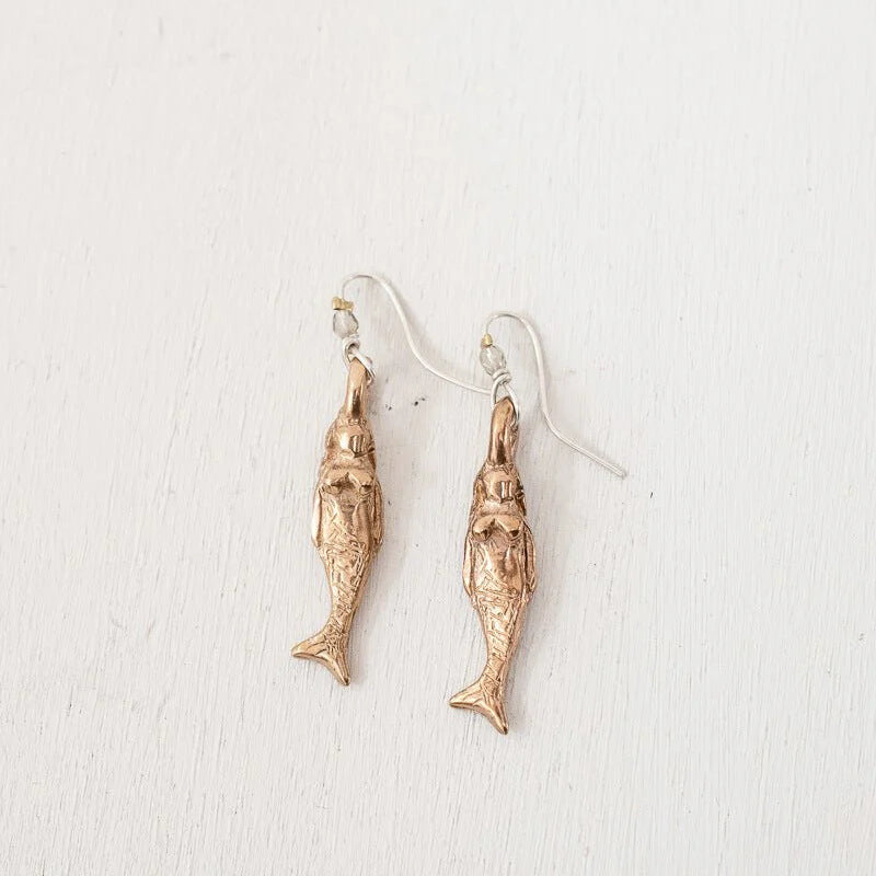A pair of mermaid shaped dangle earrings in a textured bronze finish, suspended from simple silver-toned fishhook ear wires, displayed against a clean, off-white textured background.
