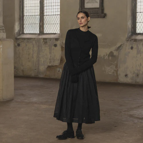 Woman in a black outfit standing in an old, dimly lit room with large windows.